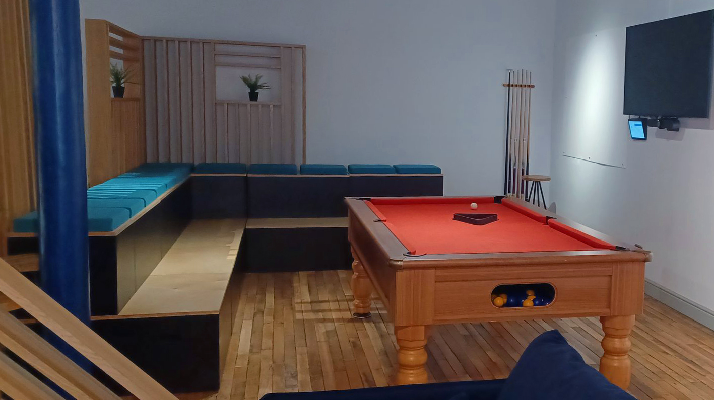 An office based pool room, with a burnt orange surface is surrounded by banked seating with comfortable teal cushions and untreated wooden dressings.