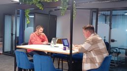 A clean, modern, attractive workspace with various members of staff busying themselves on laptops. The room is punctuated with bold colours, greenery and exposed brick walls.