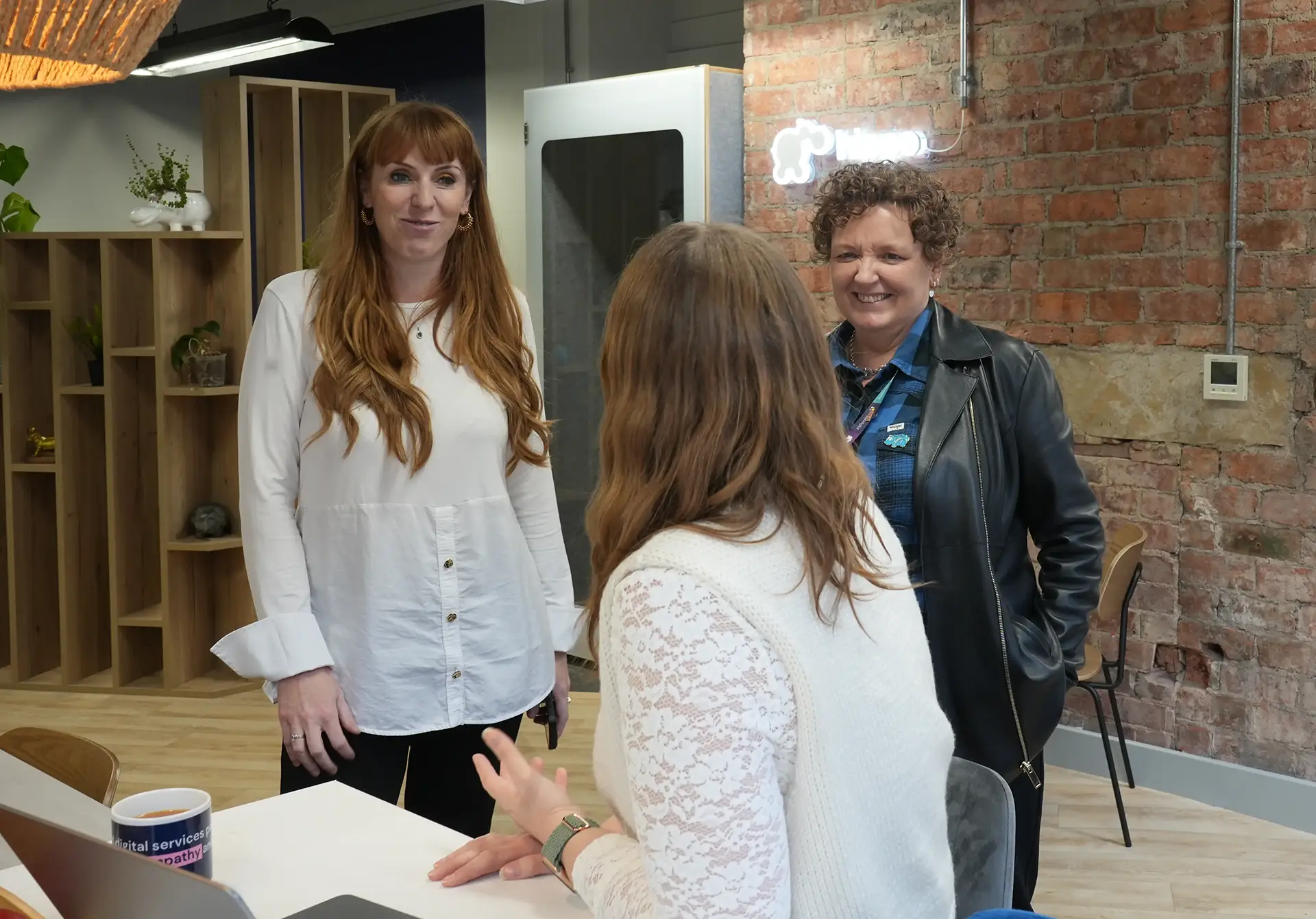 Three women stand in a modern office space, enjoying a conversation. One lady has her back to use while the other two are facing the camera engaged in conversation with the other.